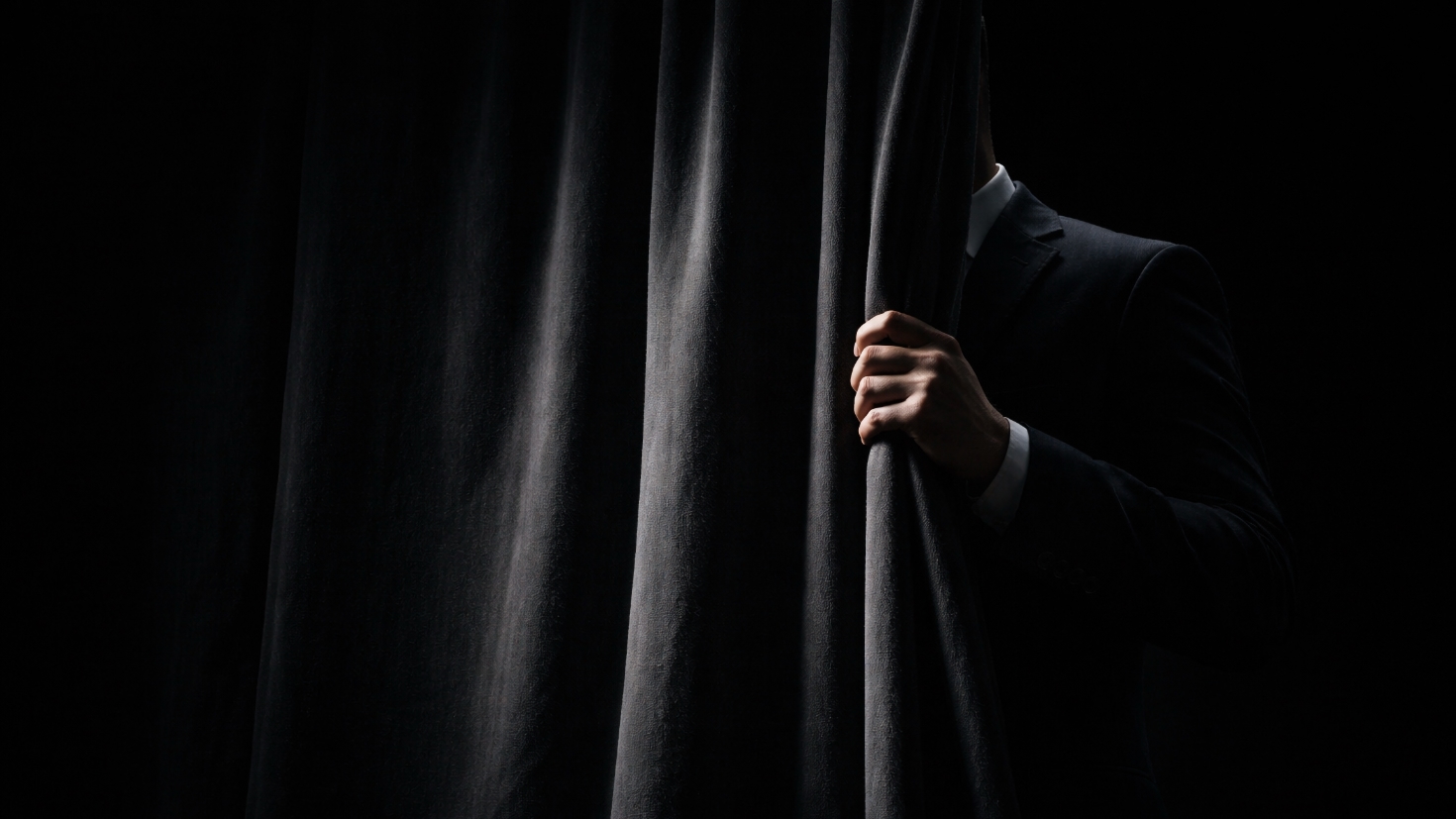 A figure in a dark suit, partially concealed behind a heavy charcoal velvet curtain, one hand gripping the curtain edge in sharp directional light against a black background — a visual metaphor for the unseen operator whose workings a director is expected to trust without seeing. (Image generated by ChatGPT 5.4) A figure in a dark suit, partially concealed behind a heavy charcoal velvet curtain, one hand gripping the curtain edge in sharp directional light against a black background — a visual metaphor for the unseen operator whose workings a director is expected to trust without seeing. (Image generated by ChatGPT 5.4)